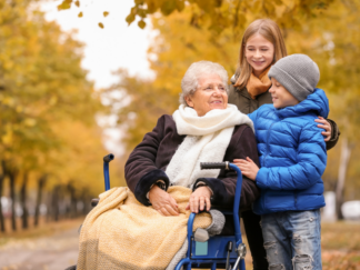 An elderly woman in a wheelchair in the foreground surrounded by two younger children in a park setting with autumnal coloured trees and leaves in the background.