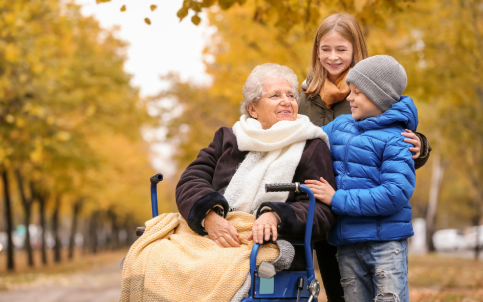 An elderly woman in a wheelchair in the foreground surrounded by two younger children in a park setting with autumnal coloured trees and leaves in the background.